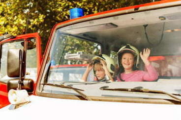 Dwoje dzieci siedzi w kaskach w samochodzie strażackim w kaskach i macha przez przednią szybę.